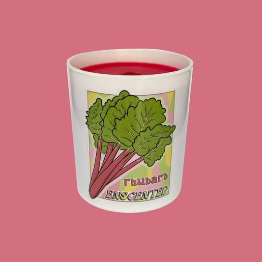 A white candle with a red tint on top, featuring an illustration of rhubarb leaves and the word 'RHUBARB' on the side, set against a pink background.